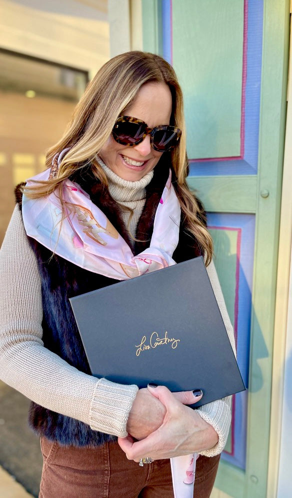 Image of beautiful woman in doorway with the Designer Shoe Print Scarf draped over shoulders and tied in front of chest, while holding the Lisa Courtney Designer box that scarf comes in.