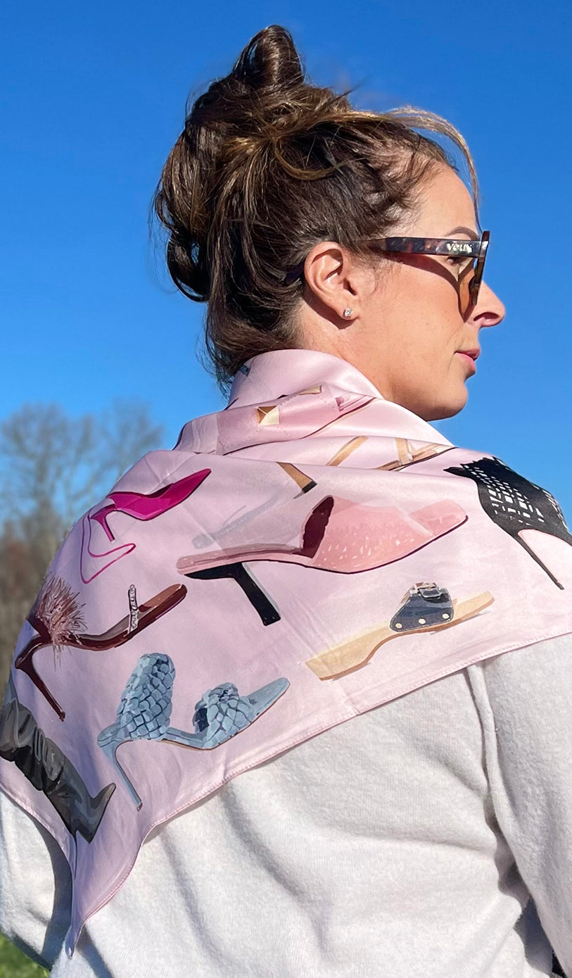 An image of a woman from behind with the Designer Shoe Scarf by Lisa Courtney Designs folded in half and draped over shoulders. Light pink silk scarf printed with colorful women’s high heels.